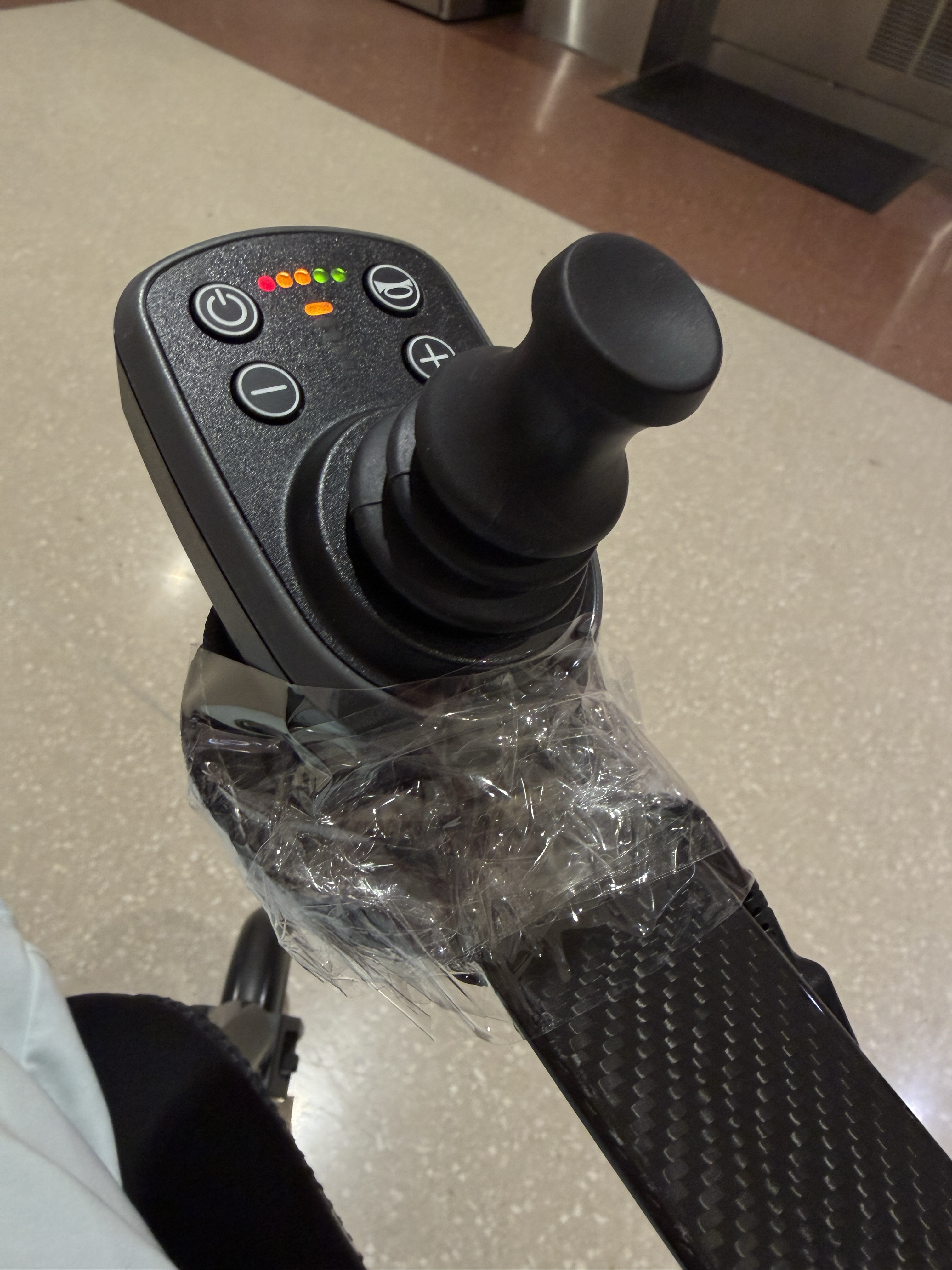 Close-up of a power wheelchair joystick controller resting on the chair's cupholder, wrapped in packing tape to hold it together after the mounting bracket snapped during the flight.