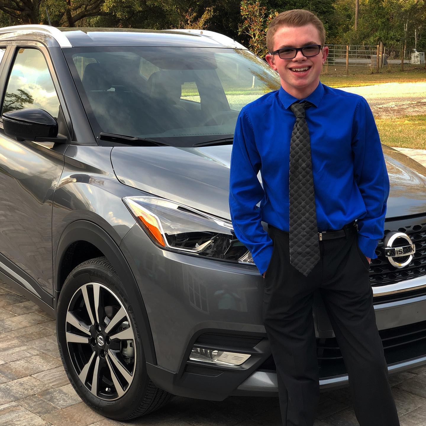 Ryleigh Newman standing beside the gray Nissan Kicks in a blue dress shirt and dark tie, hands in his pockets, smiling at the camera on a paver driveway with trees behind him.