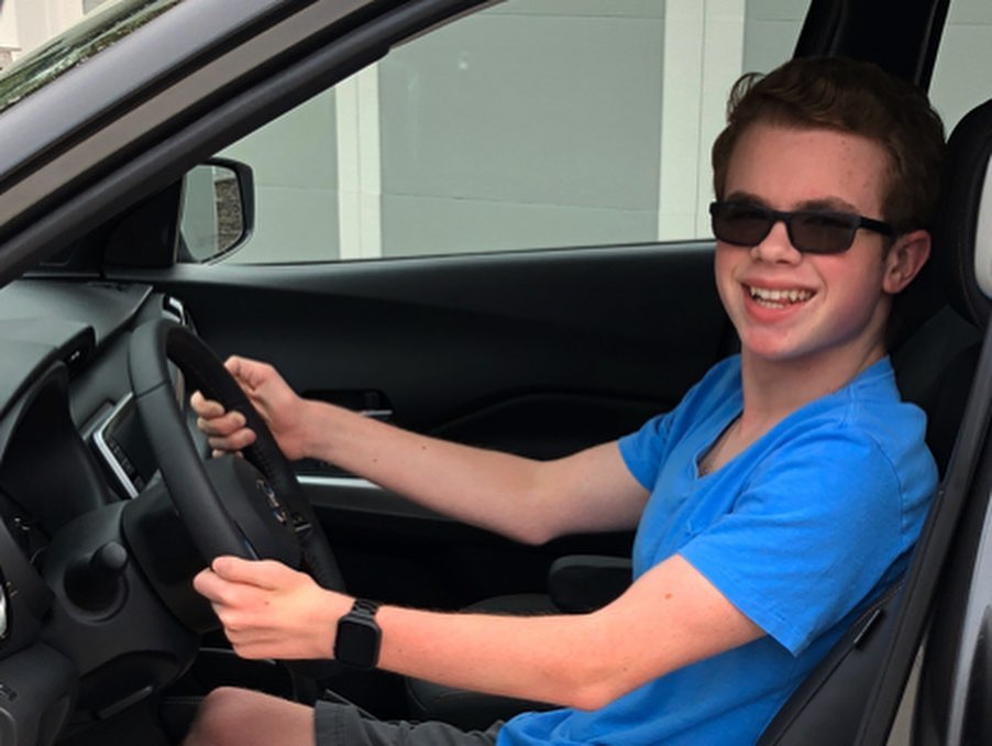 Ryleigh Newman in the driver's seat of the Kicks in a blue T-shirt and sunglasses, both hands on the steering wheel, smiling brightly at the camera through the open driver's door in a driveway.