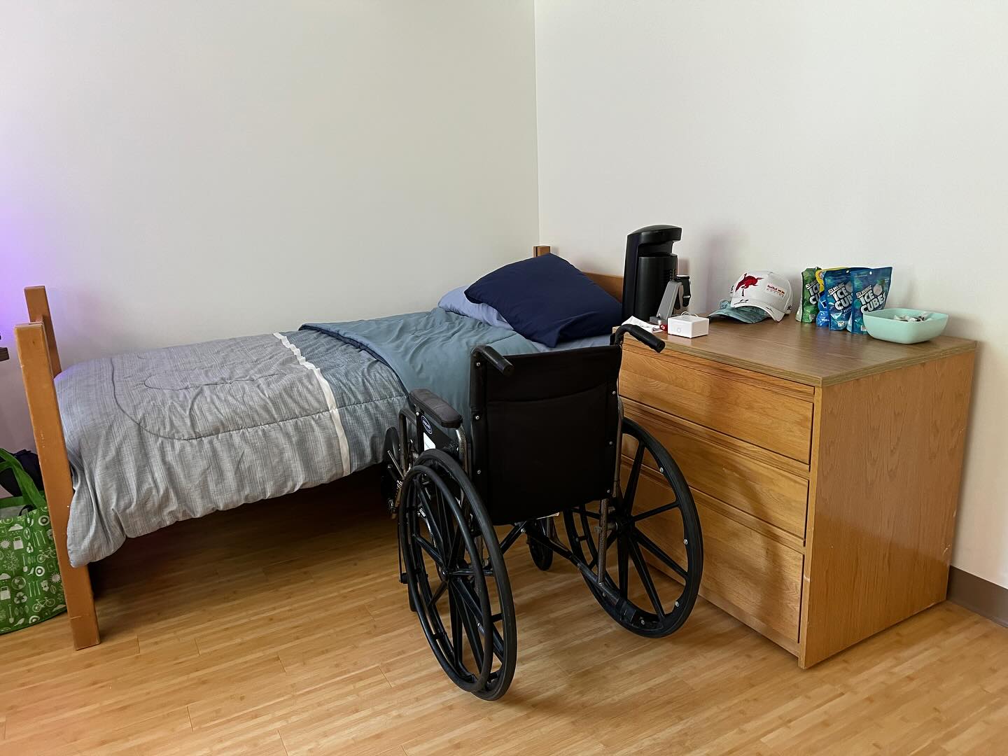 A manual wheelchair parked beside a made dorm bed with a blue comforter, next to a wooden dresser holding a coffee maker and snacks, in a bright accessible dorm room.