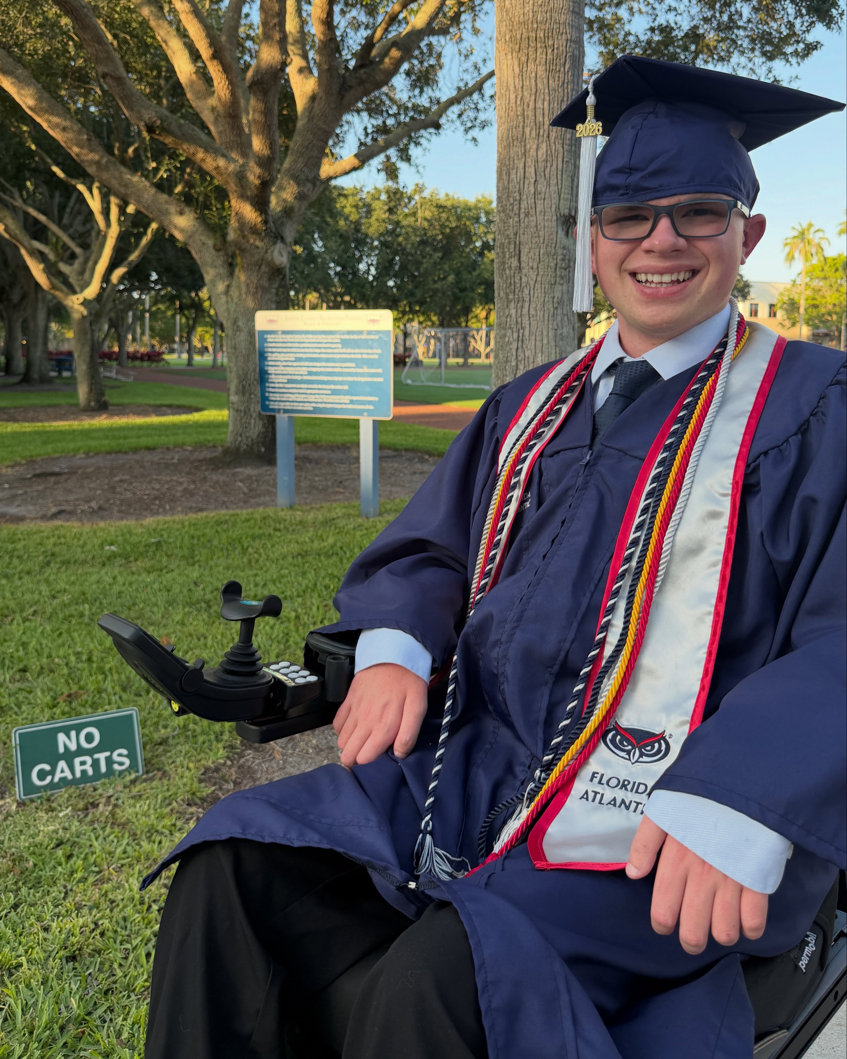 Ryleigh Newman seated in full graduation regalia under a campus tree at golden hour, honor cords and Florida Atlantic stole draped over his gown.