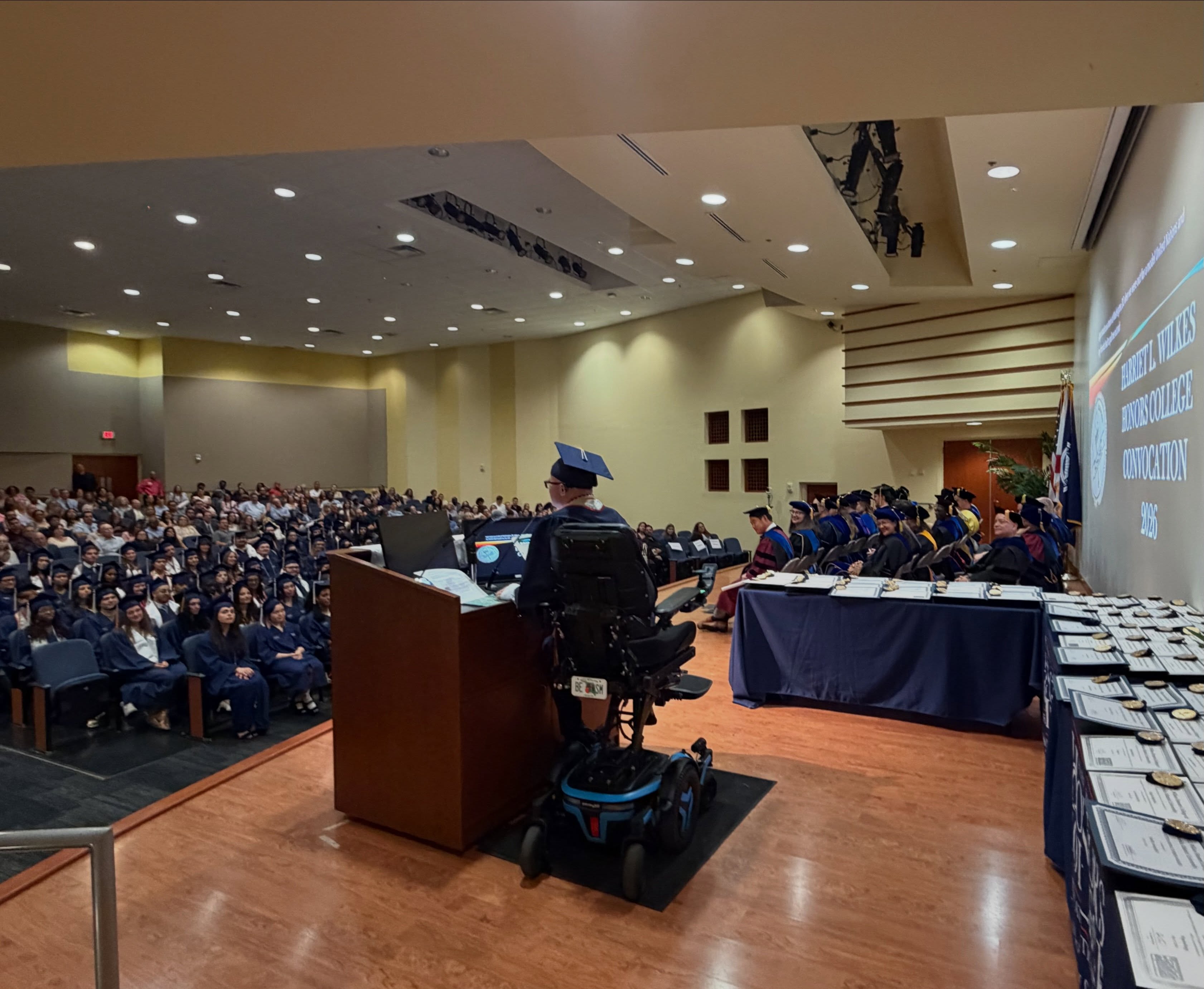 View from the stage behind Ryleigh Newman as he addresses a packed Wilkes Honors College Convocation hall, with the faculty table and rows of medallions on his right and the 2026 Convocation slide glowing on the wall.