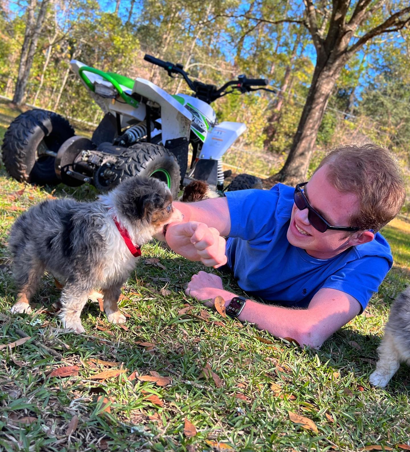 Ryleigh Newman lying on the grass in sunglasses, holding out a fist toward a small grey merle Aussie puppy who is leaning in to meet it, a small four-wheeler parked behind them.