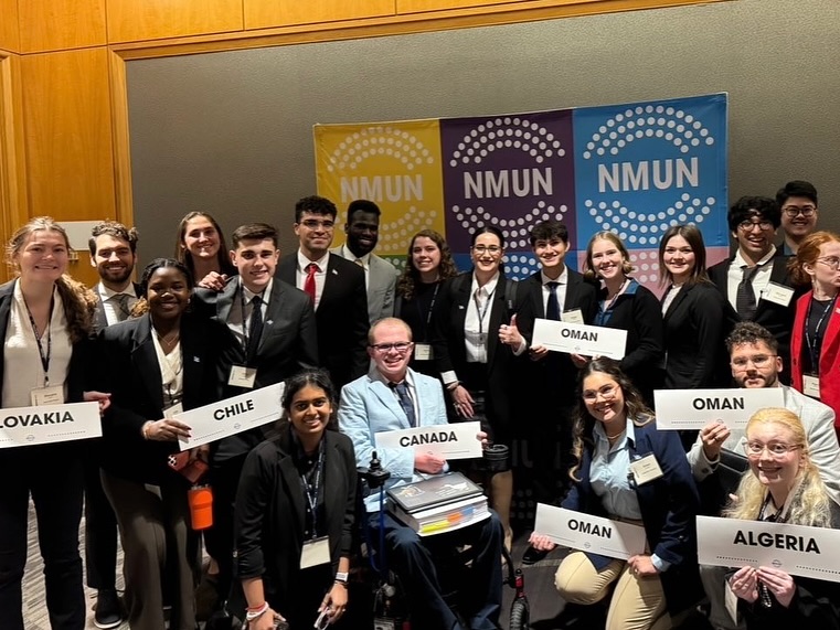 The Florida Atlantic University delegation in business attire holding country placards for Canada, Chile, Oman, Slovakia, and Algeria in front of a yellow, purple, and blue NMUN banner. Ryleigh Newman is in front holding the Canada sign.
