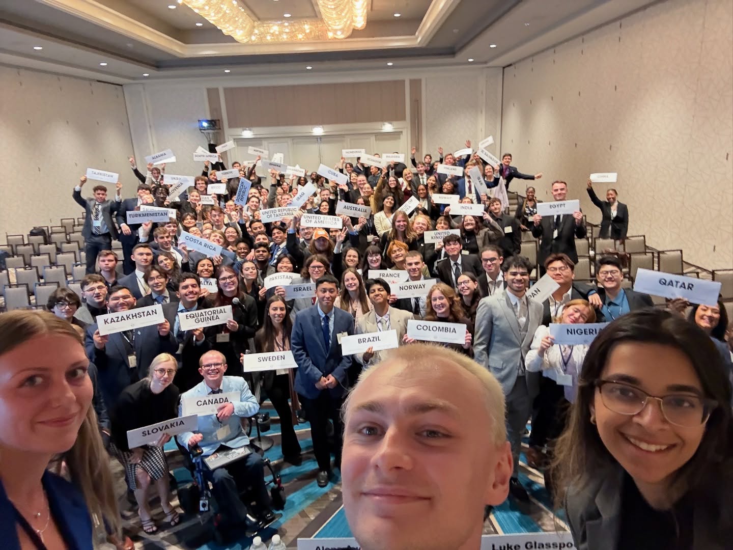 A wide selfie over a full General Assembly room of delegates holding up country placards including Kazakhstan, Equatorial Guinea, Brazil, Canada, and Qatar, with Ryleigh Newman in the foreground.