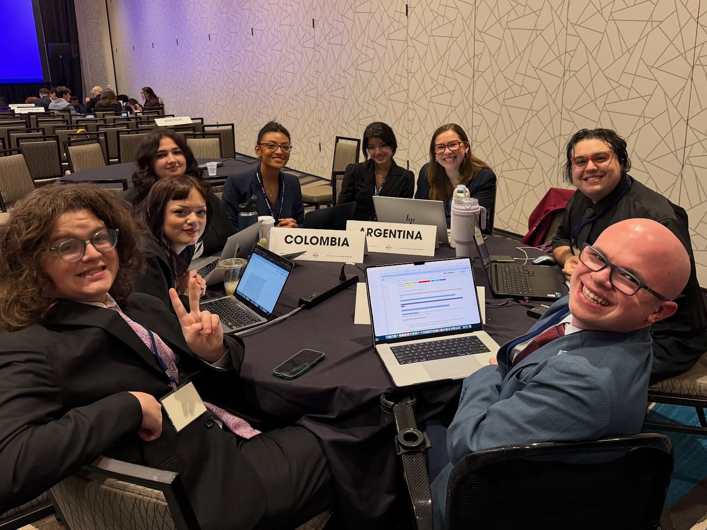 Delegates seated around a committee table covered in laptops and Colombia and Argentina placards, Ryleigh Newman smiling at the near end of the table.
