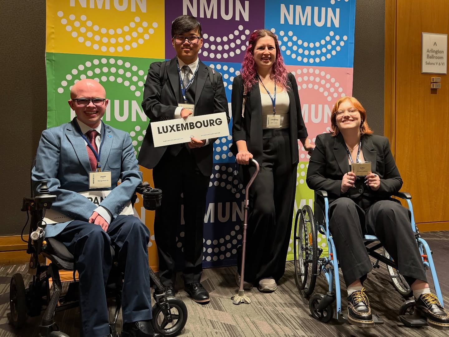 The FAU delegation posing in front of a colorful NMUN banner, several delegates holding country placards including United States of America and Australia, with Ryleigh Newman in a navy blazer on the right.
