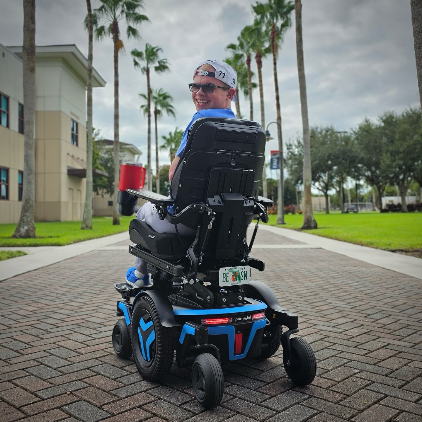Ryleigh Newman turned to smile over his shoulder while seated in his blue Permobil M3 Corpus on a brick campus path lined with palm trees, the rear of the chair and its BE AWSM Florida plate facing the camera.