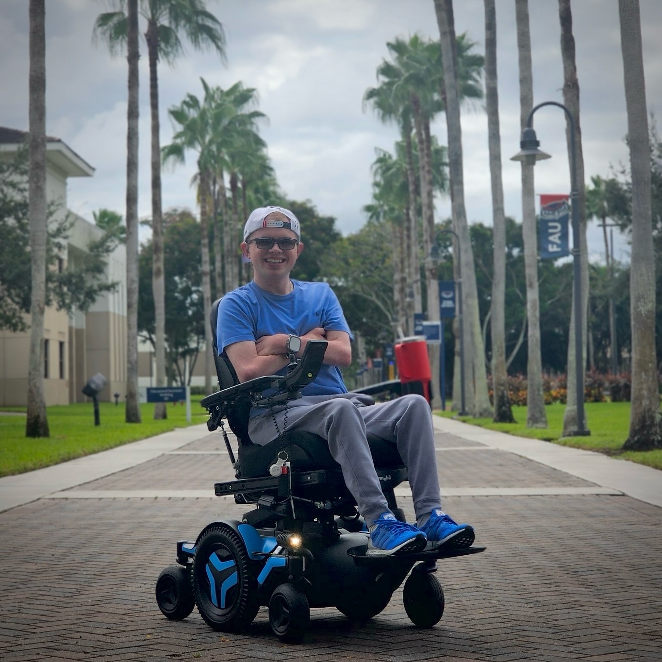 Ryleigh Newman in his Permobil M3 Corpus power wheelchair on a palm-lined campus path.