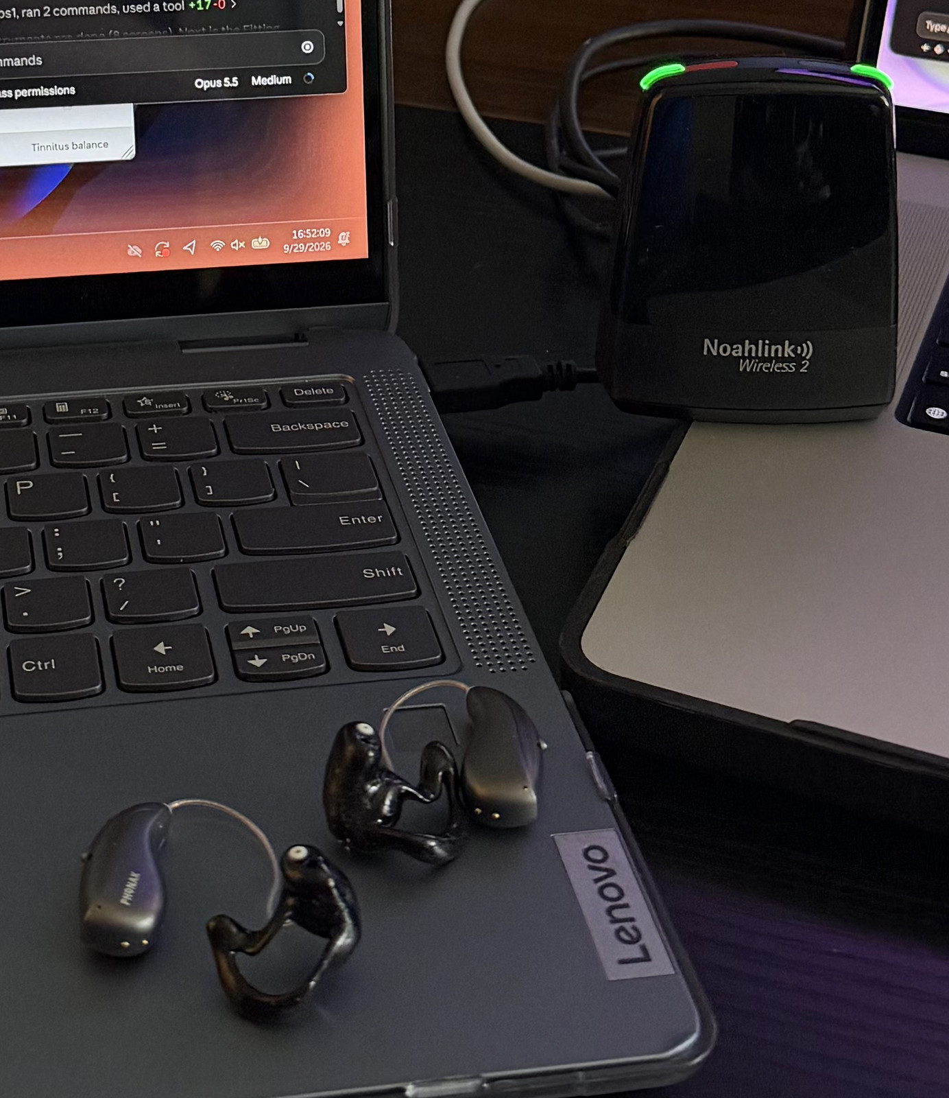 Close-up of two Phonak Audéo Sphere receiver-in-canal hearing aids with glossy black custom shells lying on a dark gray laptop, with the black Noahlink Wireless 2 programming dongle behind them, its status lights glowing green.