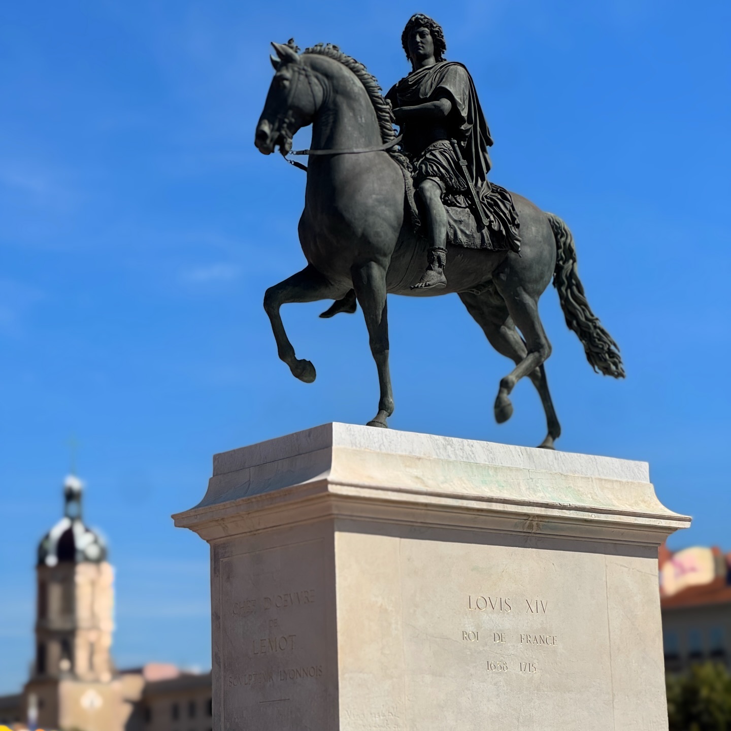 The bronze equestrian statue of Louis XIV on a tall stone plinth at Place Bellecour in Lyon, with a church bell tower in the background under a bright blue sky.