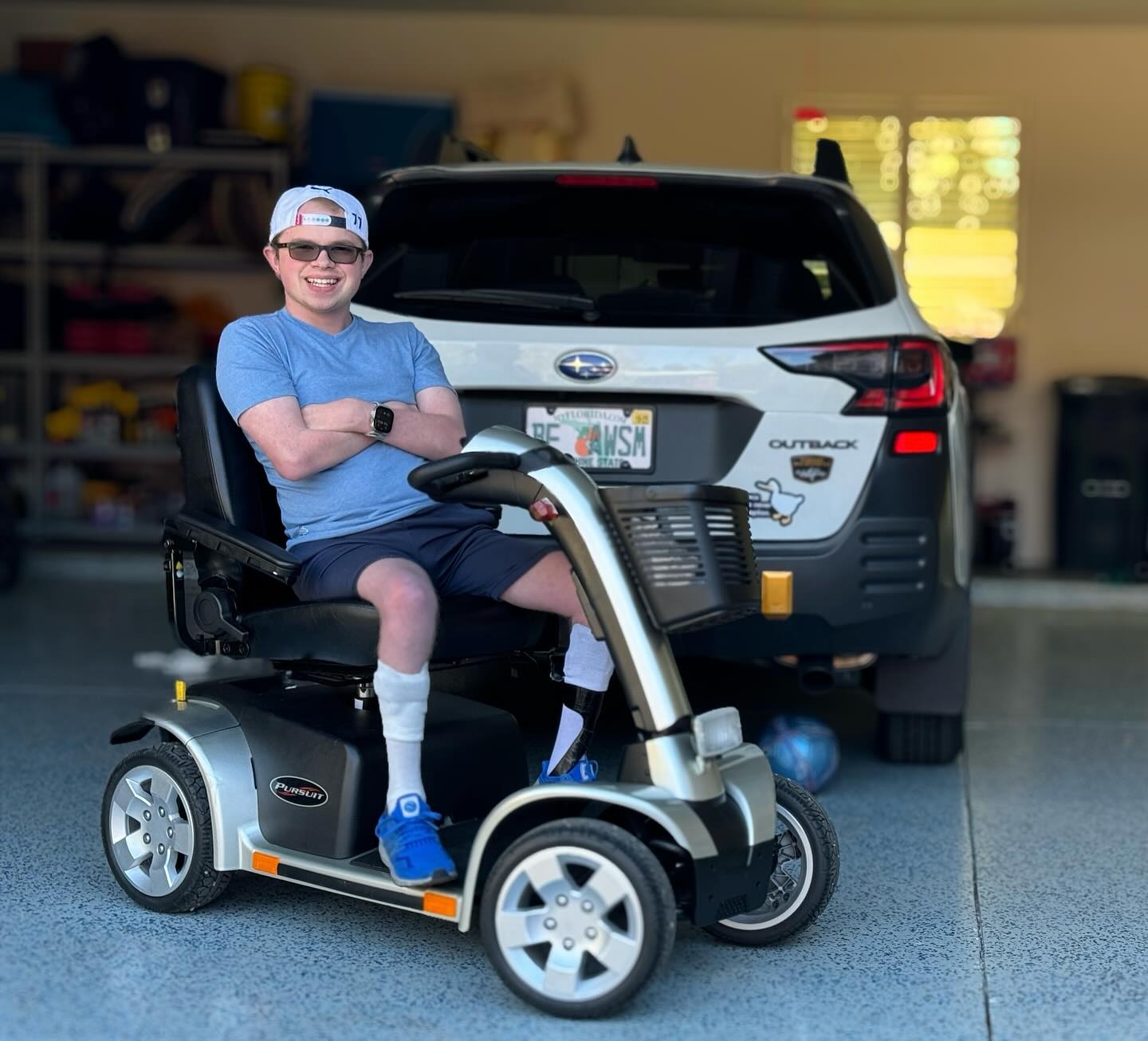 Ryleigh Newman seated on a silver Pride Pursuit four-wheel mobility scooter in a garage, next to a Subaru Outback whose Florida plate reads BE AWSM.